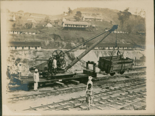 A railroad yard with workers scattered around. A wooden crane holding a cart. Single story lodges in the background on a hill in the countryside.