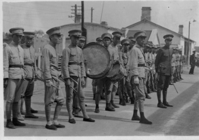 Troop of soldiers standing in a line with the leader in front of them, some soldiers have trumpets and large drums.