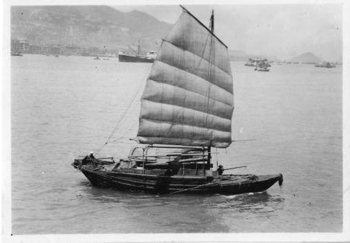 Black and white image of a Chinese sailboat called a ‘Junk’ in the calm harbor of Hong Kong. On the boat are two fishermen and further back in the waters are larger vessels (ships). The backdrop of the image is the city of Hong Kong with a towering mountain range.