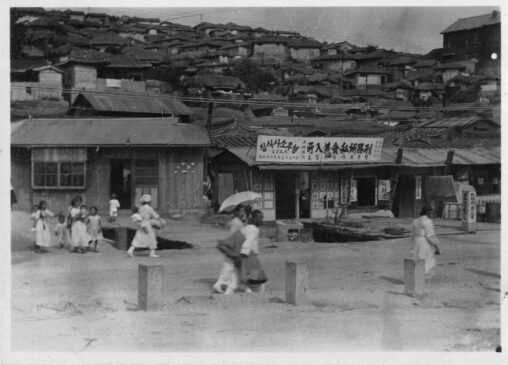 Title: Keijo, Korean Village. Caption: A photograph of many houses on a hill in Seoul, South Korea (called Keijo under Japanese rule). In the forefront, there is a business and its signs and groups of people are moving about.